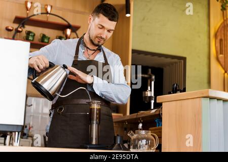 Handsome barista pours hot water in aeropress with coffee in cafe. Bearded barista works. Alternative making by barista in the cafe. Scandinavian coff Stock Photo