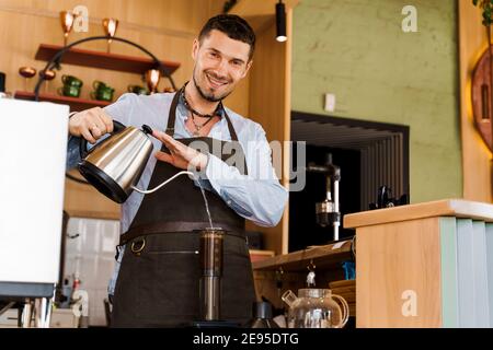 Handsome barista pours hot water in aeropress with coffee and look in camera and smile in cafe. Bearded barista works. Alternative making by barista i Stock Photo