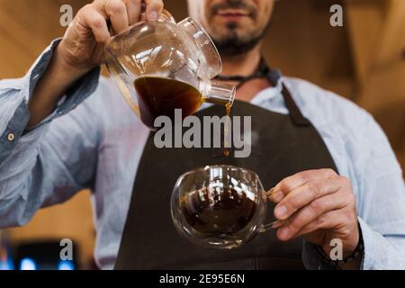 Closeup pouring coffee in double glass cup in cafe by handsome bearded barista. Coffee brewing syphon and aeropress alternative methods. Advert for so Stock Photo