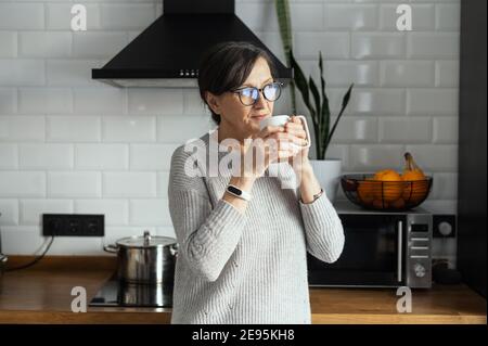 Contemporary senior woman wearing glasses enjoys morning coffee in the kitchen at home. A retirement lady daydreaming with a mug of hot drink looks Stock Photo