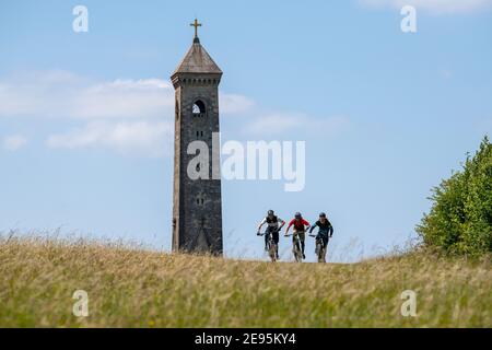 Three men ride mountain bikes past the Tyndale Monument, a tower built on a hill at North Nibley, Gloucestershire, England during the summer. Stock Photo