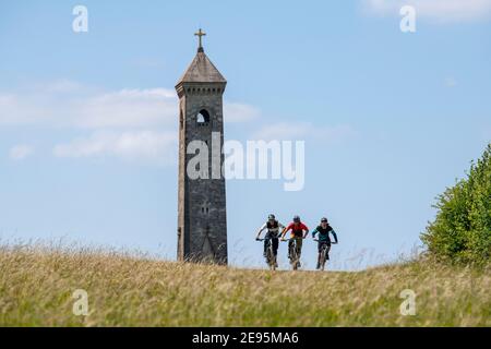 Three men ride mountain bikes past the Tyndale Monument, a tower built on a hill at North Nibley, Gloucestershire, England during the summer. Stock Photo