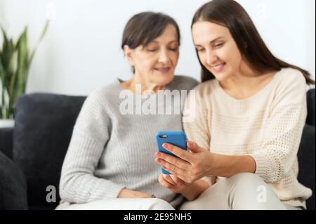 An adult daughter explains to senior mother how to send messages on the smartphone, help to register account, two multigenerational women sitting on Stock Photo