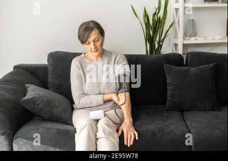 Senior woman is using tonometr for health self checking sitting on the sofa, an older lady measuring blood pressure at home. Healthcare and medicine Stock Photo