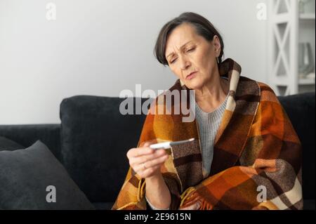 An older lady measures body temperature with a thermometer. Sick senior woman has fever, cold and flu symptoms, sits on the couch covered with a Stock Photo