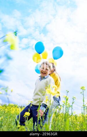 Vertical portrait happy two children run on a yellow field, blooming rapeseed. blue sky and clouds. Blue and yellow balloons, concept of freedom Stock Photo