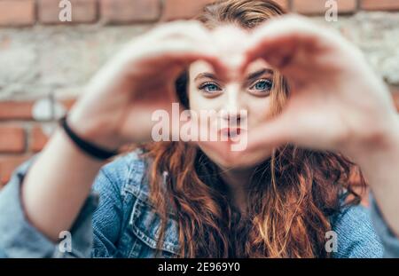 Portrait of red curled long hair caucasian teen girl with applied red lipstick lips with blue eyes on the brick wall background making a heart shape w