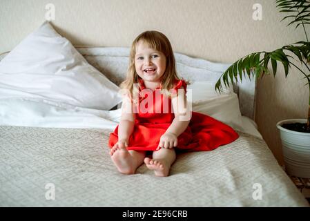 Cute red-haired blue-eyed emotional baby girl laughs. Lies on the bed in the bedroom, the bed is a white bed, a red dress for a child. pillows and Stock Photo