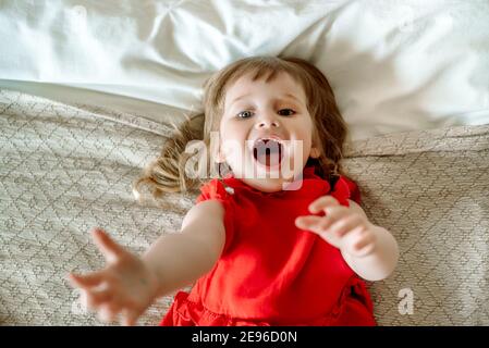 the child reaches up, cries and calls. Emotional cute baby girl in a bad mood, angry and sad. lying on the bed.asks for help, Top View Stock Photo