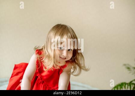 emotional beautiful baby girl jumping on the bed in a bright bedroom. In a red dress. Homeliness. White pillows, green plants. emotion of joy. Stock Photo