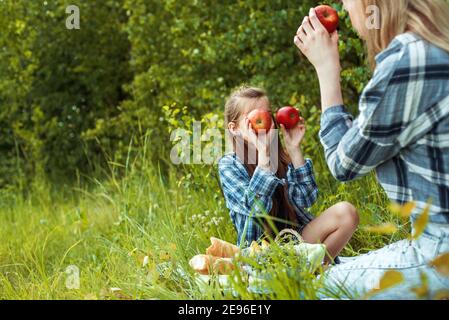 Mother with daughter in the park. mother holding the red apples near the eyes.summer park, fruit basket,white bread. outdoors. Family holidays and Stock Photo