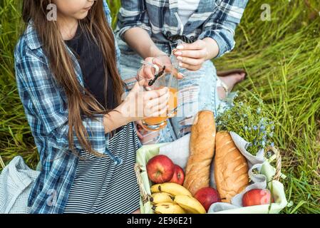 young mother and little daughters in bright sweaters on picnic in nature indulge.Picnic in the forest, green park. wicker basket, fruit, juice and Stock Photo
