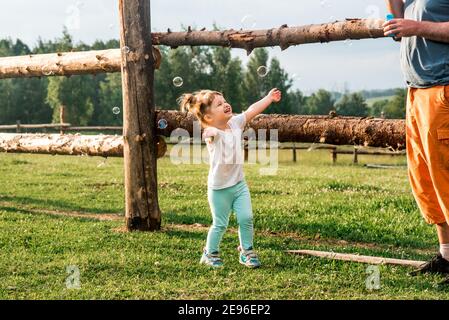 dad and daughter are making bubbles in the park.Cheerful beautiful baby girl runs and laughs, catches bubbles. happy childhood. Sunny summer evening, Stock Photo