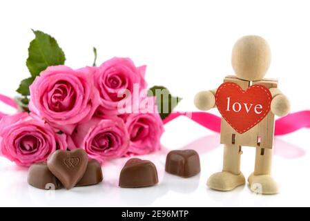 Heart-shaped chocolates, pink roses and wooden man standing and holding red heart over white background. Valentine's day concept, selective focus. Stock Photo