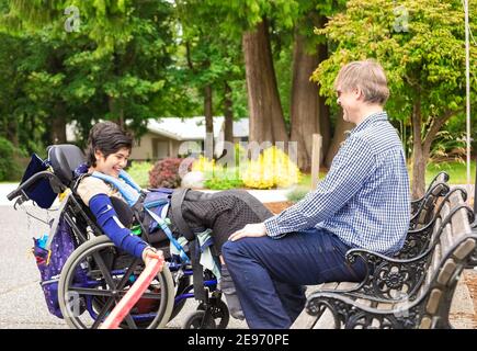 Caucasian father enjoying talk with biracial disabled young son in wheelchair while sitting on park bench, laughing together Stock Photo