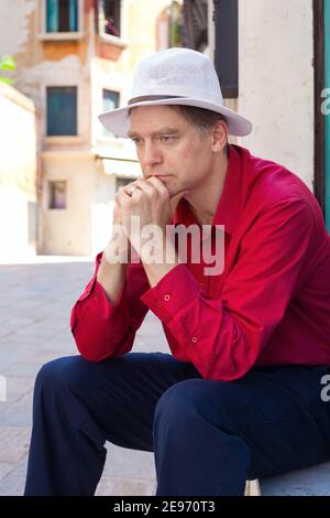 Caucasian man in red shirt and white hat with worried expression sitting outdoors in Italy Stock Photo