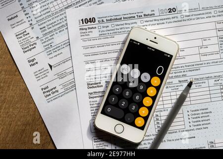 The form 1040, U.S. individual income tax return for the year 2020, an iPhone showing the calculator app screen, and a pen are seen on a desk. Stock Photo