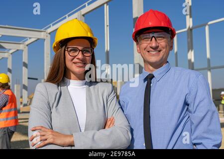 Man and Woman In Working Helmets Standing and Looking at Camera on Site. Business, Building and Teamwork Concept. Stock Photo