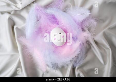 Diy for kids. Do it yourself. Funny painted smiley egg face in bird feather. Easter decorations. Copy space for text. Pastel colors. Easter greeting card. Stock Photo