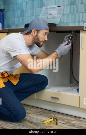 Fixing leaks. Focused young repairman, professional plumber wearing tool belt crouching on the floor while fixing sink pipe in the kitchen indoors Stock Photo