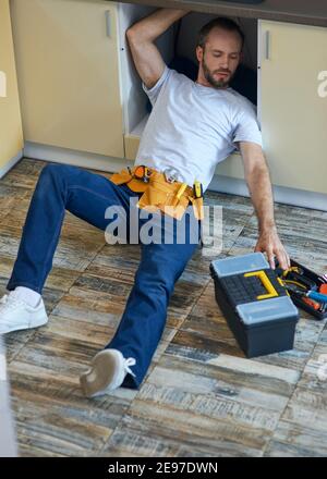 Plumbing expert. Professional repairman, plumber wearing tool belt examining and fixing sink pipe in the kitchen indoors Stock Photo