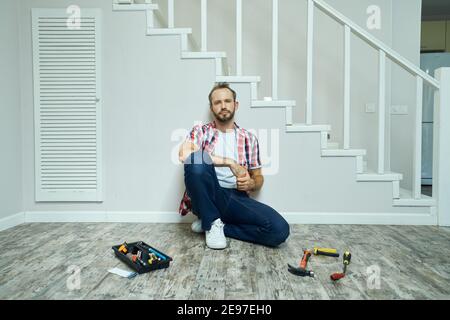 Tired handyman looking at camera, taking a break while sitting on the floor in apartment. Home repair hand tools on laminate flooring Stock Photo
