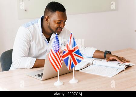 English and British flags in front. Black handsome translator uses his laptop for online work according social distancing. Scientist works at home on Stock Photo