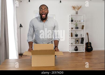 Excited African-American guy screams happily unpacking long awaited parcel box. A multiracial customer overjoyed with postal shipping Stock Photo
