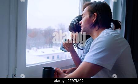 Happy multiracial couple drinking morning coffee and looking hrough the window at the park covered in snow. High quality photo Stock Photo