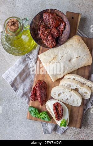 Sliced ciabatta bread with sun-dried tomatoes, cheese, basil and olive oil on wooden board, stone background. Selective focus. Stock Photo