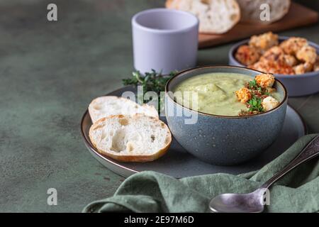 Broccoli cream soup decorated with thyme, cream, roasted cauliflower and fresh bread on green concrete background. Healthy vegetarian food concept. Se Stock Photo