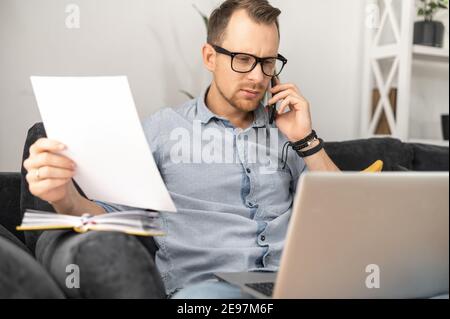 An intelligent busy businessman wearing eyeglasses working on laptop from home, talking on smartphone and holding documents, doing paperwork, checking Stock Photo