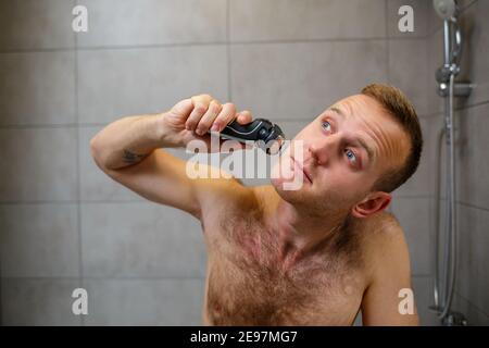 A man shaves his face with an electric razor in front of a mirror. Skin irritation. Bath procedure Stock Photo