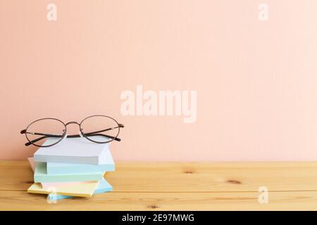 Glasses and stack of colorful memo pad, sticky notes on wooden desk. Pink background, copy space. Work and study place Stock Photo