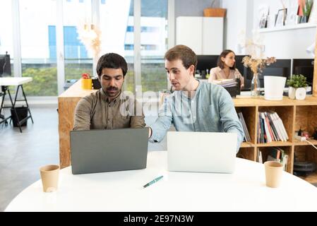Two male coworkers working on their laptops helping each other. Stock Photo