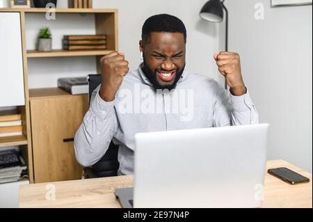 Happy excited African-American businessman celebrating his success, winner of the competition raising hands up, expressing joy from achieving goals Stock Photo