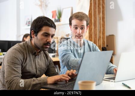 Coworkers helping each other while working on their laptops in the office. Stock Photo
