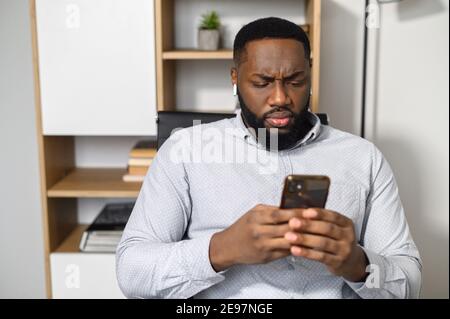 Pensive puzzled African-American man in smart casual shirt holding smartphone and expressing misunderstanding, texting message, answering email indoor Stock Photo