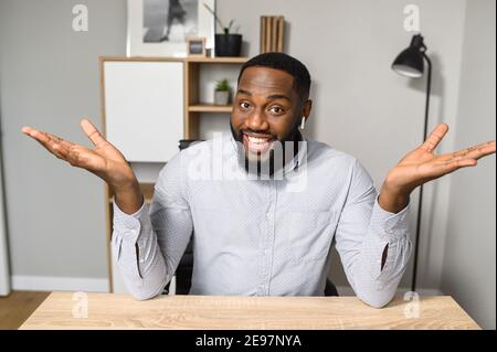 Excited cheerful young African-American guy sitting at the desk and raising hands wide open, smiling and looking at the camera, unexpected good news Stock Photo