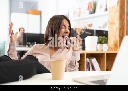 An angry hispanic woman whith her feet on desk yelling on the phone in the office. Stock Photo