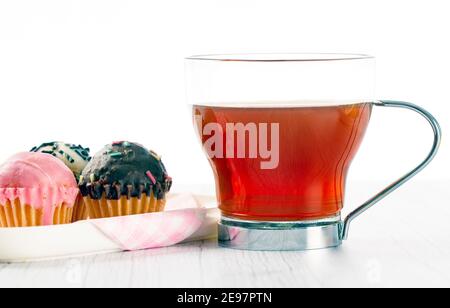 infusion on wooden surface in light tone and white background, accompanied by a plate with mini cup cakes covered in strawberry, dark chocolate and wh Stock Photo