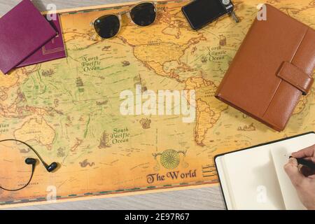 top view of a traveler's hand taking notes in his notebook to prepare for his next trip. On the table there are passports, sunglasses, headphones to l Stock Photo