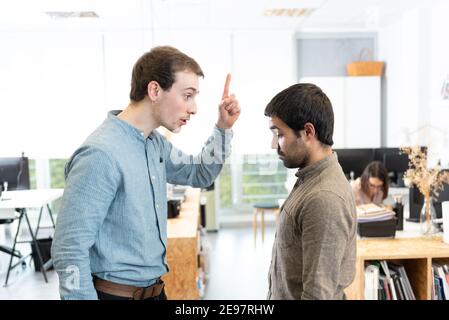 Handsome young man threatening his  sad hispanic coworker at the office. Stock Photo