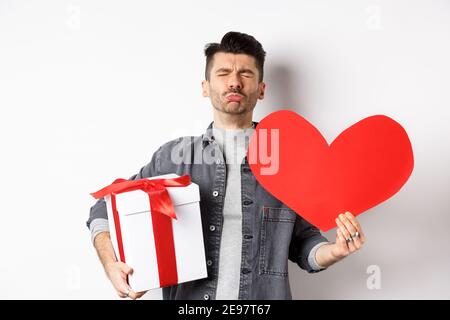 Sad and heartbroken man being rejected, crying and holding red heart with gift box, breakup on Valentines day, white background. Love and relationship Stock Photo