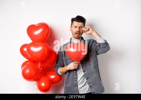 Valentines day and love concept. Sad crying man holding red heart balloon and whiping tears, standing single and miserable, being heartbroken, white Stock Photo