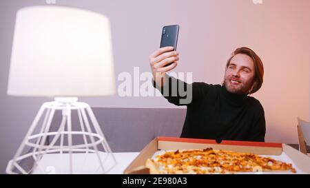 Young caucasian man having video call on his smartphone while having pizza for dinner. High quality photo Stock Photo