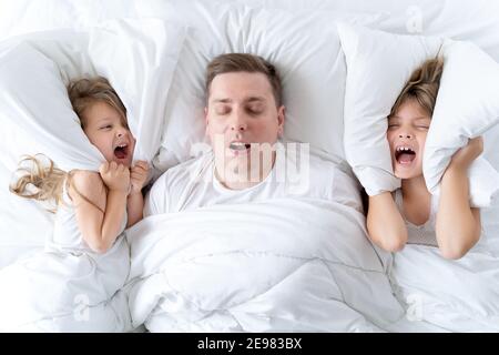 Kids, dad sleep together in bed. Father is snoring hard, girls, daughters are covering ears with pillow. Disorders, joint sleeping. Insomnia, night Stock Photo