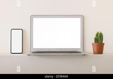 Smartphone and laptop with white blank screens placed with potted cactus on shelf against light wall Stock Photo