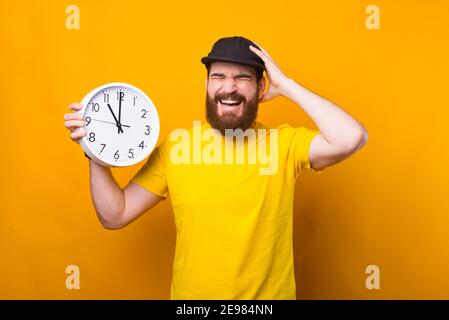 Photo of bearded young man holding wall clock and making frustrating gesture, I am late. Stock Photo