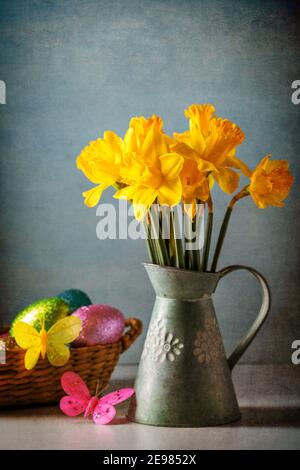Rustic styled still life of bunch of yellow daffodils in vintage water jug with Easter decorations. Selective focus, textured effect Stock Photo